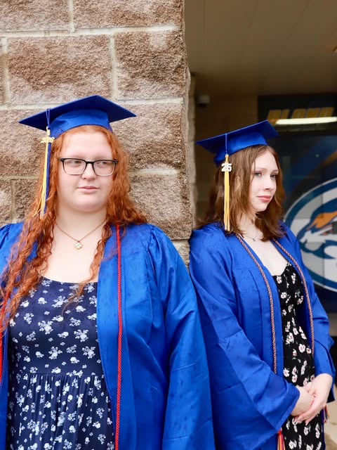 Two graduates in blue gowns and caps standing by a stone wall.
